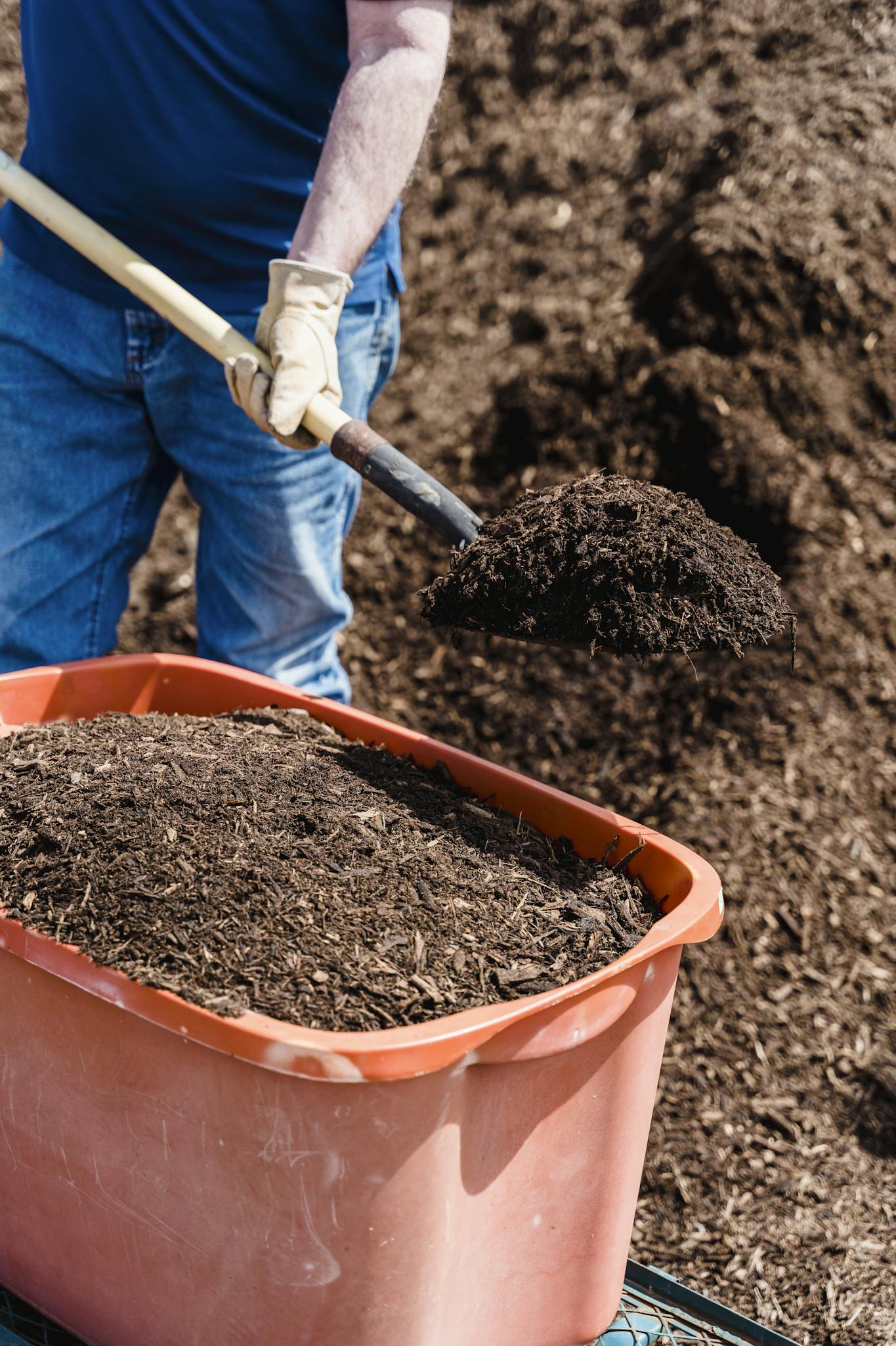 Man shoveling rich soil into a large orange compost bin in a garden.